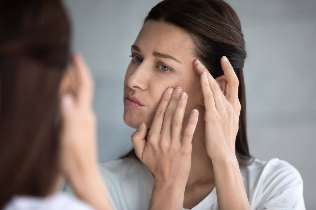 Woman closely examining her face in mirror for volume changes and contour loss after significant weight loss.
