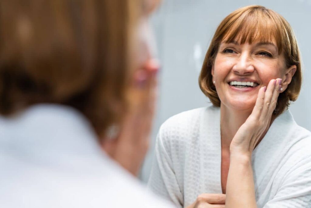 Smiling woman touching her face in the mirror, showing healthy skin before deep plane facelift preparation.