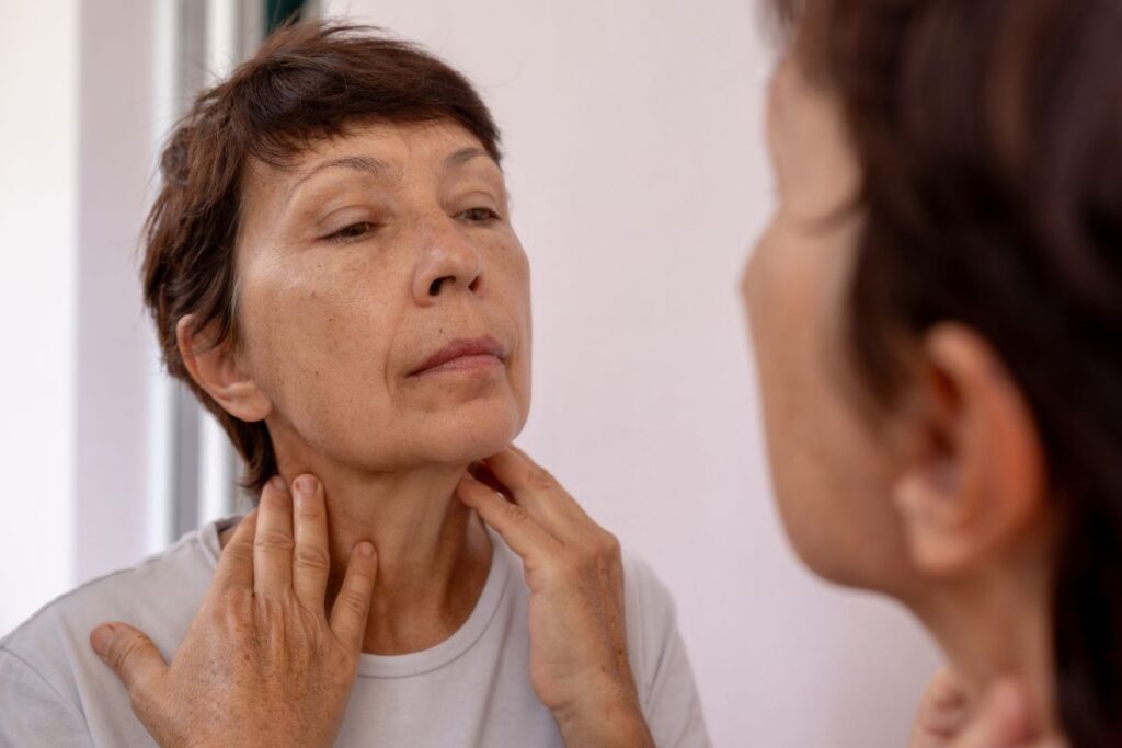 Mature woman examining neck and jawline in mirror, assessing facial volume loss and early aging signs