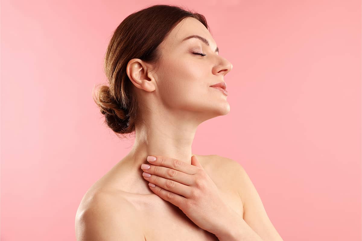 A profile view of a woman with her hair in a bun tilting her head back with eyes closed, resting her hand gently against her lower neck.