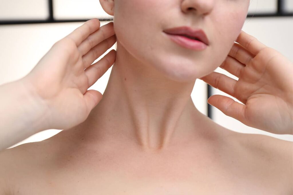 A close-up view of a woman’s neck and collarbone area as she raises both hands to frame her jawline.