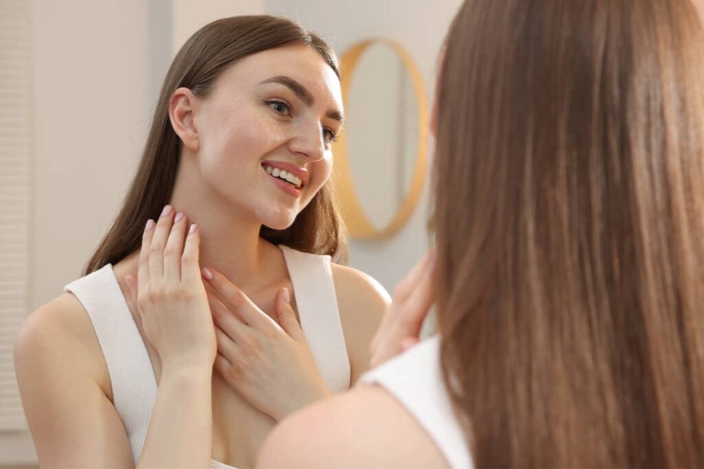 A smiling young woman looking at her reflection in a mirror while gently touching her smooth chin and neck area.