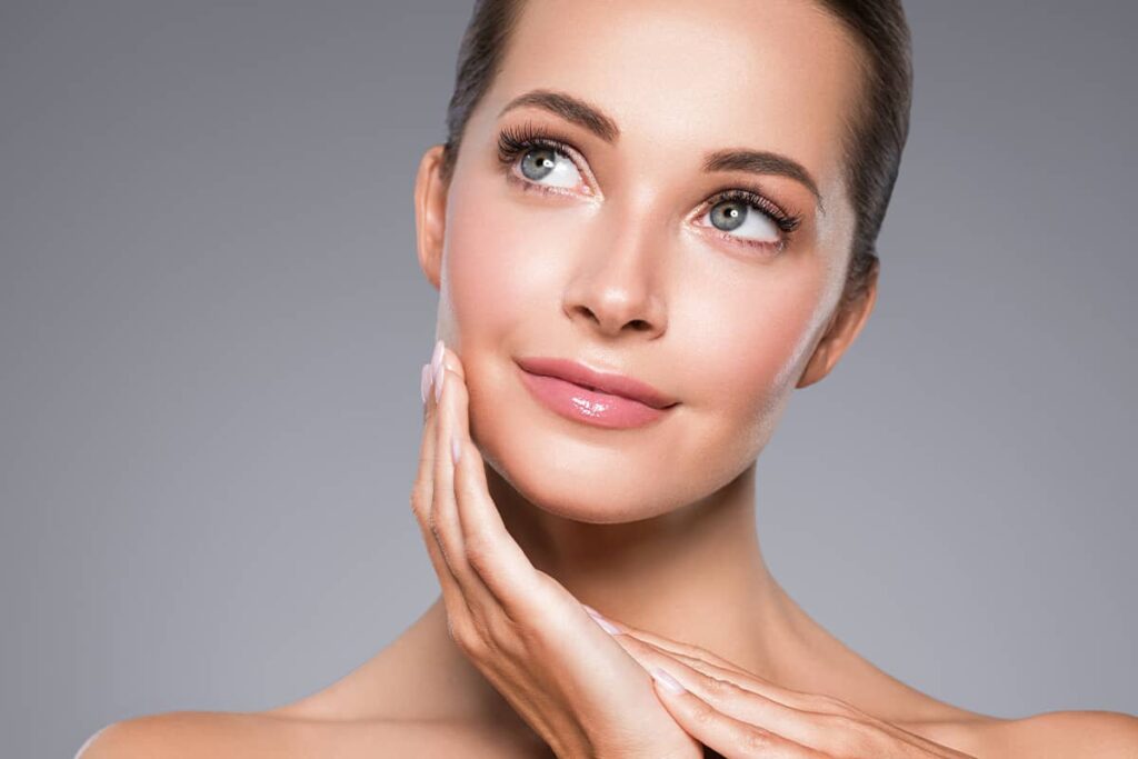 A close-up view of a smiling woman with clear, luminous skin looking up and to the side while holding her hand to her jawline.