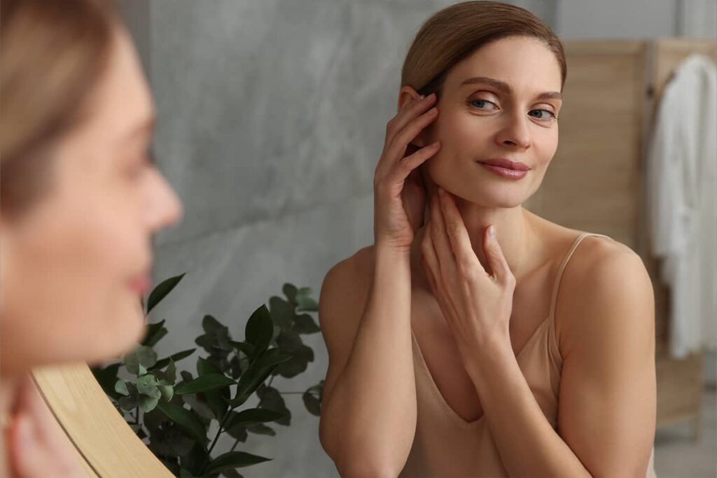 A woman looking at her reflection in a bathroom mirror while using her hands to examine the firmness of her cheek and jawline skin.